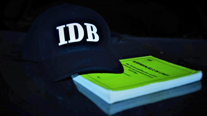 Attēls ar melnu cepuri, uz kuras ir IDB logo un zaļa gKrimināllikuma grāmatu