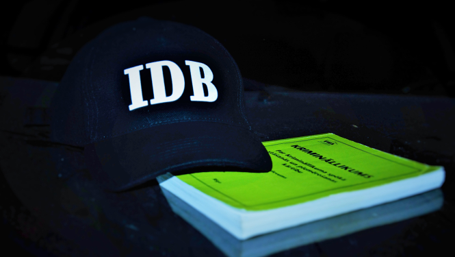 Attēls ar melnu cepuri, uz kuras ir IDB logo un zaļa gKrimināllikuma grāmatu