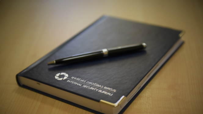 Fotogrāfija ar gadagrāmatu un pildspalvu. Uz klades ir Iekšējās drošības biroja logo