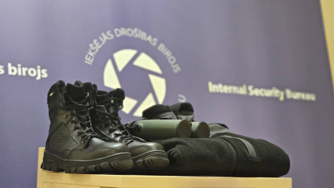 Fotogrāfija, kurā redzami melni zābaki, tālskatis zaļā krāsā un melna flīsa jaka. Fonā redzams tumši zils baneris ar Iekšējās drošības biroja logo baltā krāsā.