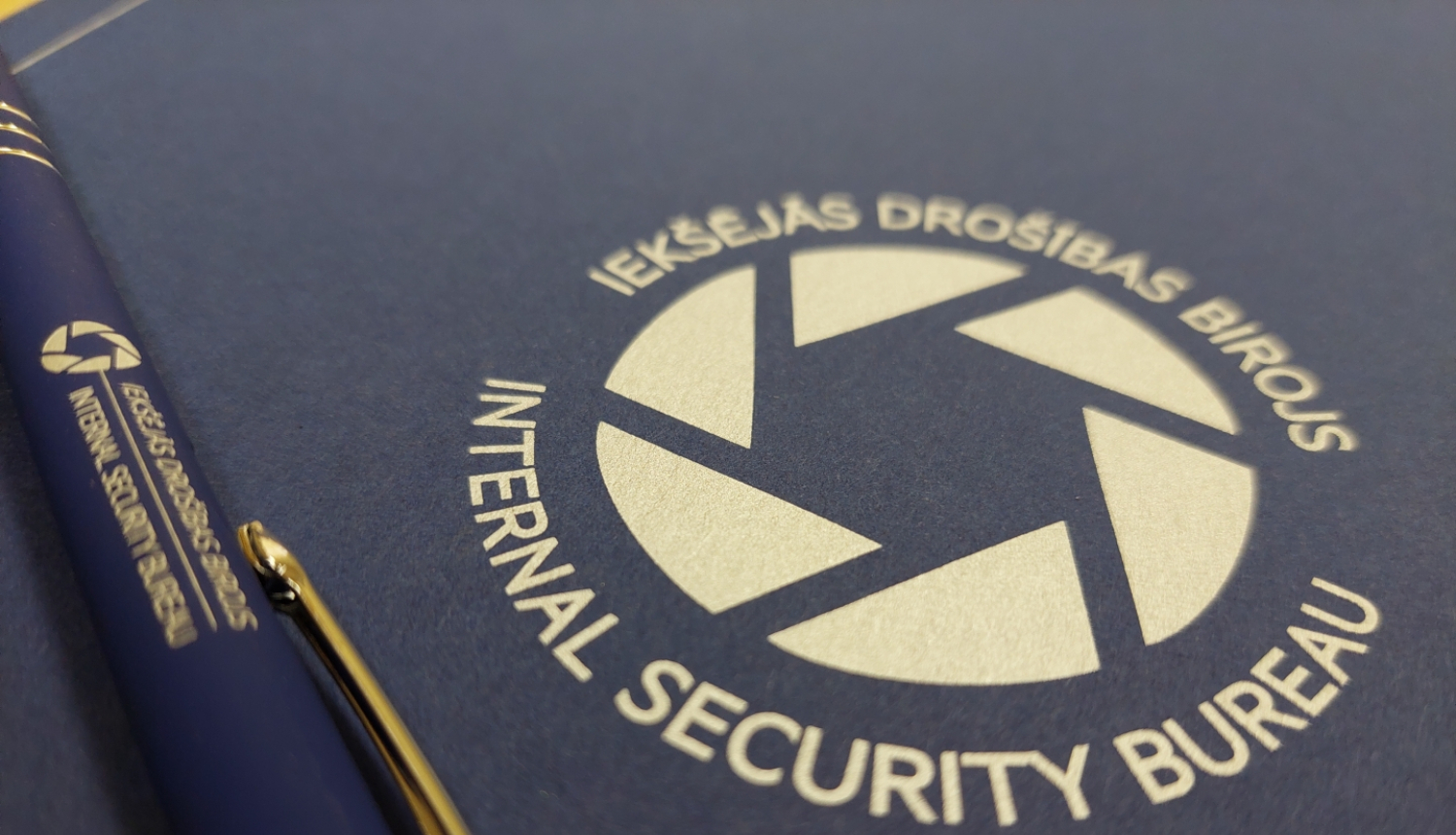 Attēls ar Iekšējās drošības biroja logo un zilu pildspalvu, uz kuras arī ir Iekšējās drošības biroja logo.