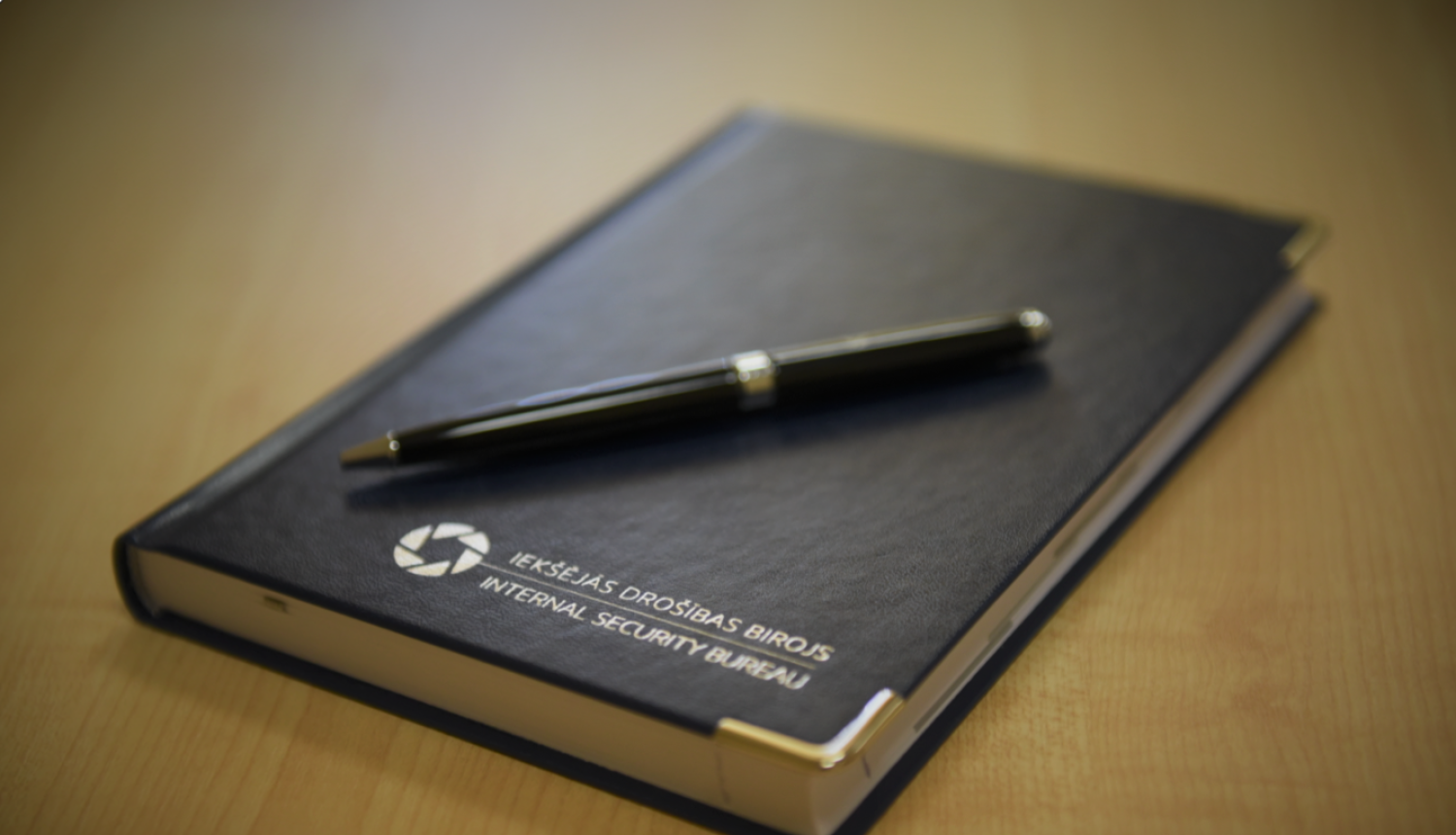 Fotogrāfija ar gadagrāmatu un pildspalvu. Uz klades ir Iekšējās drošības biroja logo