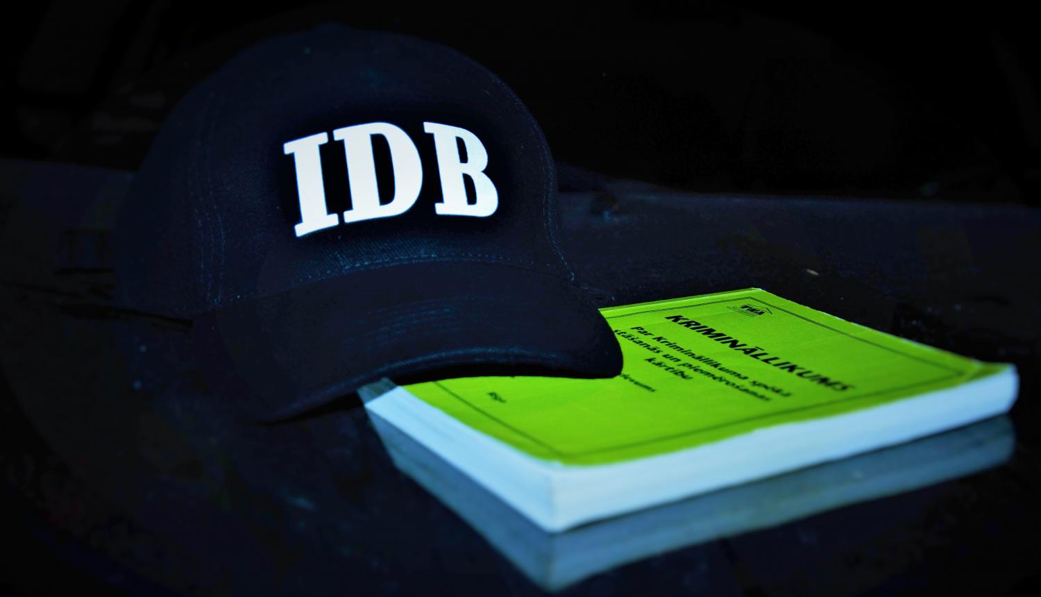 Attēls ar melnu cepuri, uz kuras ir IDB logo un zaļa gKrimināllikuma grāmatu