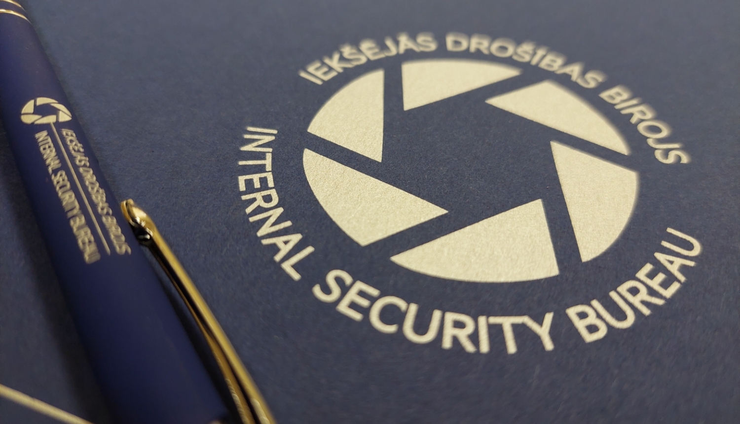 Attēls ar Iekšējās drošības biroja logo un zilu pildspalvu, uz kuras arī ir Iekšējās drošības biroja logo.